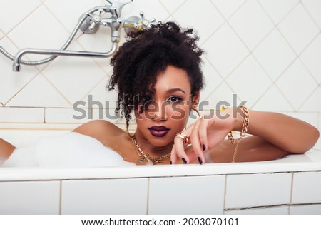 young afro-american teen girl laying in bath with foam, wearing swag jewelry flawless, making selfie