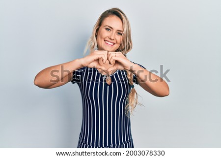 Beautiful young blonde woman wearing casual striped dress smiling in love doing heart symbol shape with hands. romantic concept. 