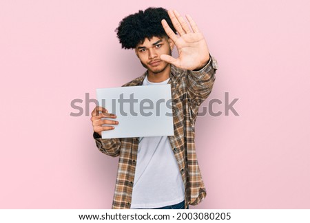 Young african american man with afro hair holding blank empty banner with open hand doing stop sign with serious and confident expression, defense gesture 