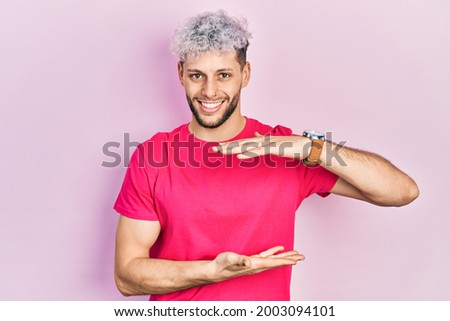 Young hispanic man with modern dyed hair wearing casual pink t shirt gesturing with hands showing big and large size sign, measure symbol. smiling looking at the camera. measuring concept. 