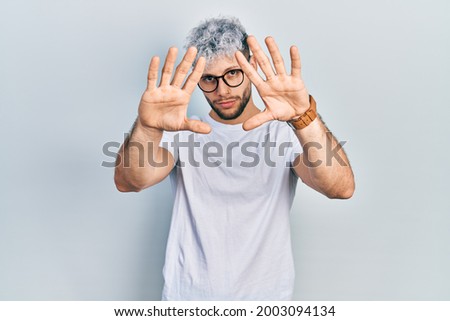 Young hispanic man with modern dyed hair wearing white t shirt and glasses doing frame using hands palms and fingers, camera perspective 