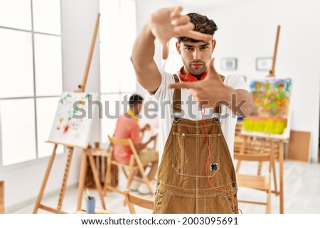 Young hispanic man at art studio smiling making frame with hands and fingers with happy face. creativity and photography concept. 
