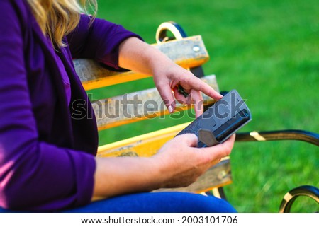 Defocus close-up female hands searching on the phone outside, outdoor. Woman in purple blouse using smartphone while sitting on a bench in the park. Copy space. Green nature background. Out of focus.