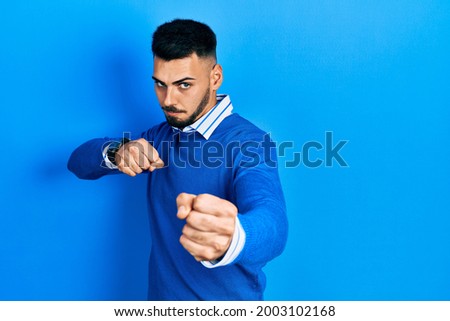 Young hispanic man with beard wearing casual blue sweater punching fist to fight, aggressive and angry attack, threat and violence 