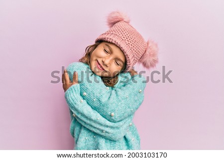 Little beautiful girl wearing wool sweater and cute winter hat hugging oneself happy and positive, smiling confident. self love and self care  Royalty-Free Stock Photo #2003103170