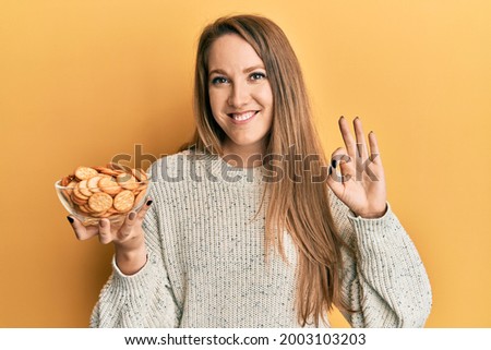 Young blonde woman holding salty biscuits bowl doing ok sign with fingers, smiling friendly gesturing excellent symbol 