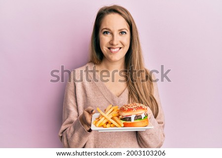 Young blonde woman eating a tasty classic burger with fries smiling with a happy and cool smile on face. showing teeth. 