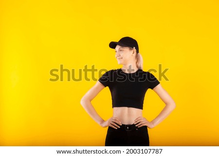 Joyfull girl in black slothes smiling looking aside on yellow background. Pretty blond blue-eyed girl model in black high waist jeans, black short t-shirt and black cap posing.