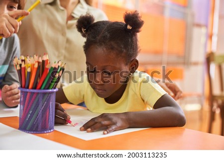 Creative African girl with pigtails paints picture with crayons in painting class in kindergarten