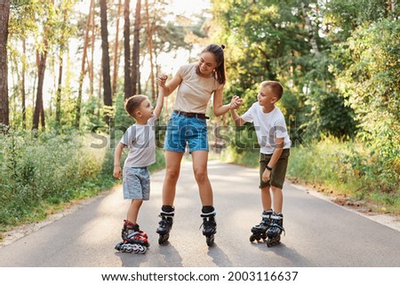 Young mother with her children rollerblading in summer park, holding arms her sons and smiling happily, spending time and having fun together, family wearing casual clothing.
