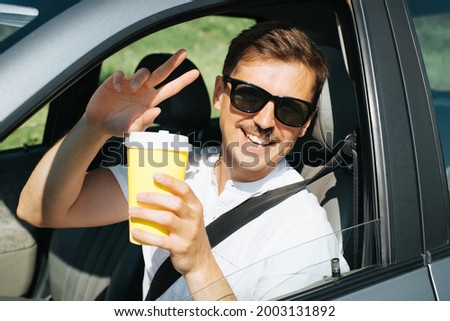 Joyful young man in sunglasses holding a disposable cup of coffee while driving, attractive driver sitting in the car and showing the peace sign through the open window. Summer road trip