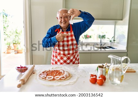 Senior man with grey hair cooking pizza at home kitchen smiling making frame with hands and fingers with happy face. creativity and photography concept. 
