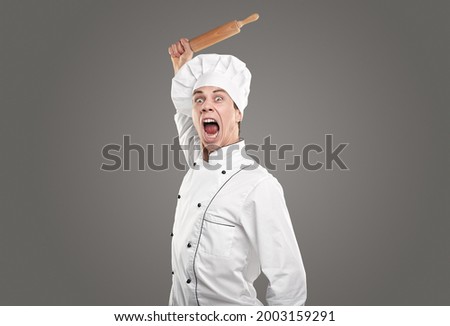 Insane male cook in white chef uniform and hat holding wooden rolling pin in raised hand and screaming loudly against gray background