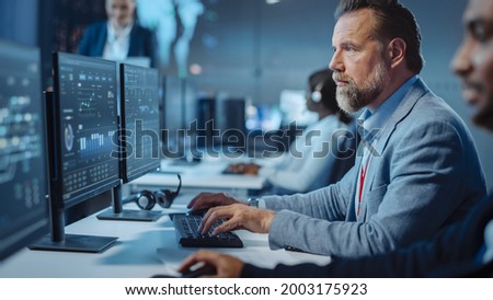 Portrait of Serious Professional Technical Controller Sitting at His Desk with Multiple Computer Displays Before Him. In the Background His Colleagues Working in System Control and Monitoring Center.