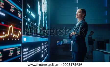 Young Female Computer Science Engineer Looking at Big Screen Display Showing Global Map with Data Points. Telecommunications Technology Company System Control and Monitoring Room with Servers. Royalty-Free Stock Photo #2003175989
