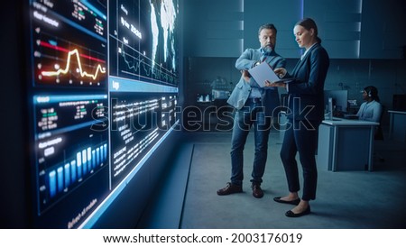 Project Manager and Computer Science Engineer Talk while Using Big Screen Display and a Laptop, Showing Infrastructure Infographics Data. Telecommunications Company System Control and Monitoring Room. Royalty-Free Stock Photo #2003176019