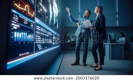 Project Manager and Computer Science Engineer Talk while Using Big Screen Display and a Laptop, Showing Infrastructure Infographics Data. Telecommunications Company System Control and Monitoring Room. Royalty-Free Stock Photo #2003176028