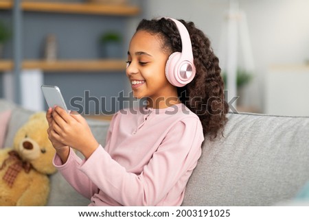 Music application. Joyful african american girl listening favourite songs in wireless headphones and chatting on her smartphone, scrolling social media feed while sitting on sofa at home and smiling