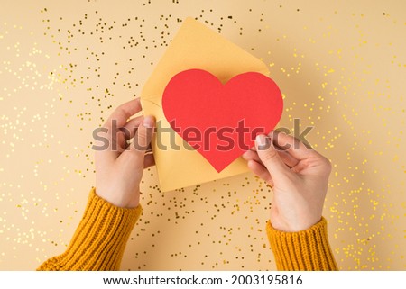 First person top view photo of female hands in yellow sweater holding open pastel yellow envelope with red paper heart over scattered golden sequins on isolated pastel orange background with copyspace Royalty-Free Stock Photo #2003195816