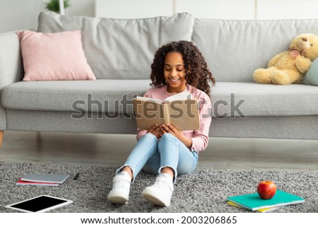 Youth and literature. Smiling african american gen z girl holding and reading paper book. Schoolgirl sitting on the floor carpet at home in cozy living room, leaning on couch Royalty-Free Stock Photo #2003206865