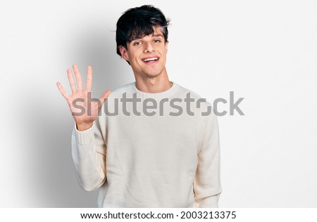 Handsome hipster young man wearing casual winter sweater showing and pointing up with fingers number five while smiling confident and happy. 