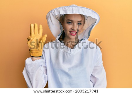 Beautiful blonde caucasian woman wearing protective beekeeper uniform showing and pointing up with fingers number three while smiling confident and happy. 