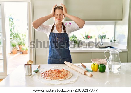 Beautiful blonde woman wearing apron cooking pizza suffering from headache desperate and stressed because pain and migraine. hands on head.  Royalty-Free Stock Photo #2003217977