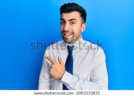 Young hispanic man wearing business clothes cheerful with a smile of face pointing with hand and finger up to the side with happy and natural expression on face 