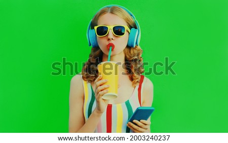 Summer colorful portrait of stylish young woman drinking fresh juice listening to music in headphones with smartphone on green background