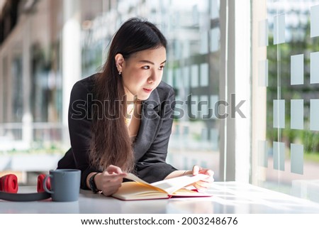 Asian businesswoman sitting by the window of a coffee shop holding a book headphones and a coffee mug at the table.