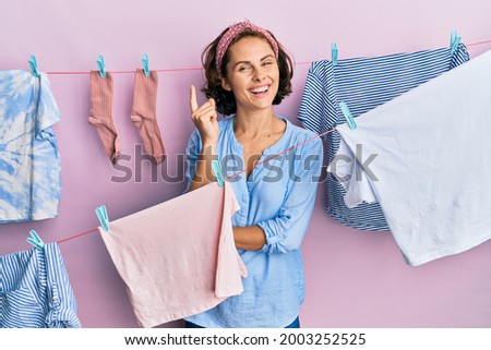 Young brunette woman doing laundry around string hangs with a big smile on face, pointing with hand and finger to the side looking at the camera. 