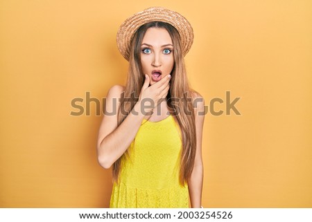 Young blonde girl wearing summer hat looking fascinated with disbelief, surprise and amazed expression with hands on chin 