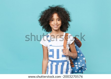 Smiling little african american kid schoolgirl 12-13 years old in striped clothes with backpack isolated on blue wall background studio. Childhood education in school concept. Mock up copy space Royalty-Free Stock Photo #2003270576