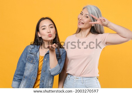 Pretty funny family asian women girls gray-haired mother brunette daughter in casual clothes posing blowing sending air kiss showing victory sign isolated on yellow color background studio portrait