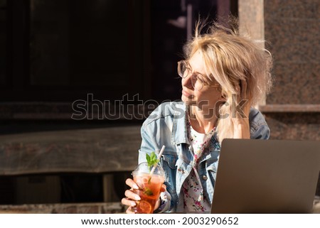a beautiful woman in glasses and a denim jacket sits thoughtfully at a table in a cafe with a laptop on the table on a summer day. Horizontal photo