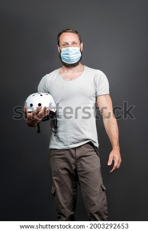 A handsome American bearded man looks into the camera and Holds a white bicycle helmet In His Hand. Brutal Russian biker in a surgical mask. Courier, delivery service, lockdown Covid-19 concept