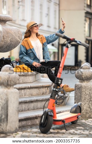 Young woman, sitting on staircase of old historical building in city center, taking picture with her phone. Female traveler exploring Ljubljana's old town on electric scooter. Summer leisure.