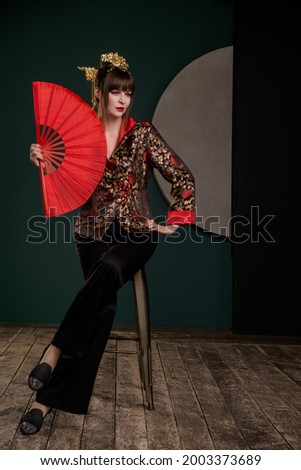 Beautiful woman with bright make up posing in Chinese costume in minimalistic bright color interior with red fan, big dagger and astrological calendar