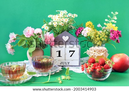  Calendar for July 17 : the name of the month of July in English, cubes with the number 17, bouquets of wild flowers, jam, fruit, cups of tea, white napkins, green background