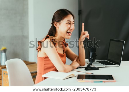 Portrait of cheerful young Asian female blogger using mic and laptop with digital tablet to interview guest during live stream at home