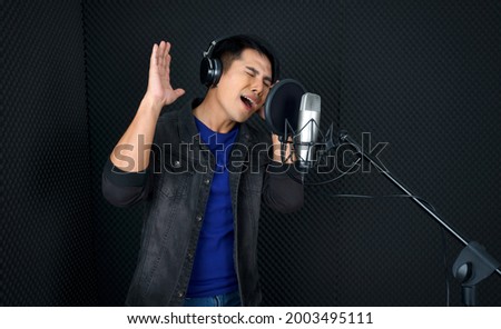 Young asian man with headphones singing in front of black soundproofing wall. Musician producing music in professional recording studio.