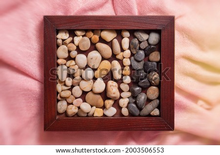A wooden frame with sea pebbles on a pink fabric. Stones of two colors-light and dark. Empty space for text, inscriptions. Photo taken from above. On the right side there are sun glare.