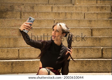 Young and beautiful girl with pigtails and punk style sitting on some stairs taking a selfie with her mobile phone.