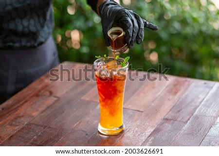 Selective focus. Clear glass. Orange juice drink with ice in the glass. And the hand of shopkeeper wearing black gloves pouring a shot of coffee into the mix. Fresh orange juice mixed with cold coffee