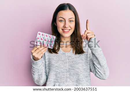 Young brunette woman holding pills smiling with an idea or question pointing finger with happy face, number one 