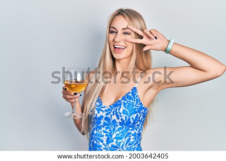 Young beautiful caucasian woman drinking a glass of white wine doing peace symbol with fingers over face, smiling cheerful showing victory 