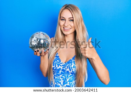Young caucasian woman holding shiny disco ball smiling happy and positive, thumb up doing excellent and approval sign 