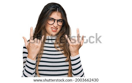 Young beautiful teen girl wearing casual clothes and glasses shouting with crazy expression doing rock symbol with hands up. music star. heavy concept. 