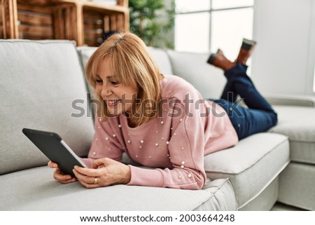 Middle age blonde woman using touchpad lying on the sofa at home.