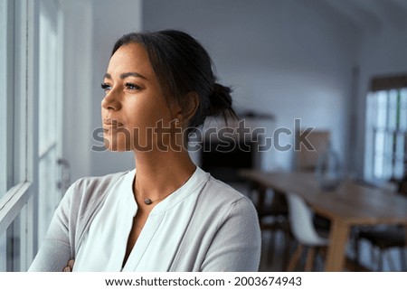 Mature african woman looking outside window with uncertainty. Thoughtful mid adult woman looking away through the window while thinking about her future business after pandemic. Doubtful lady at home. Royalty-Free Stock Photo #2003674943